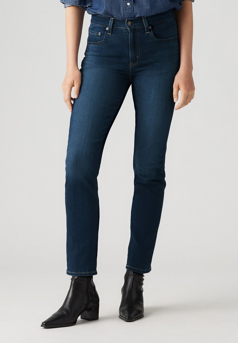 Femme portant un jean slim bleu foncé et des bottines en cuir noir, debout devant un fond gris clair uni.