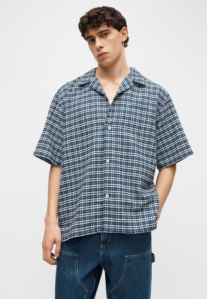 Joven con cabello corto y rizado, vistiendo una camisa azul y blanca de manga corta con cuadros y jeans azules, de pie con una mano en el bolsillo.