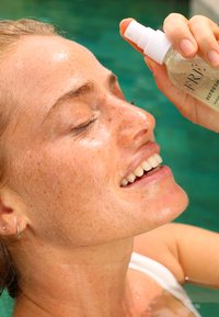 Nawilżający spray do twarzy w przezroczystej butelce z białym dozownikiem. Zbliżenie na osobę aplikującą go na twarz, ukazujące świeżą, lśniącą skórę.