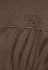 Smeđa teksturirana tkanina s glatkim gornjim dijelom i donjim dijelom u obliku vafla. Sadrži utisnuti tekst: "Home Court Advantage est 1962."