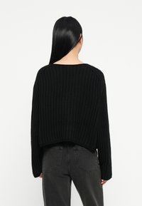 Donna con lunghi capelli neri che indossa un maglione nero a trama e jeans grigio scuro, di spalle contro uno sfondo bianco semplice.