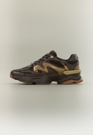 Ténis desportivo castanho escuro e dourado com sola texturizada e atacadores pretos sobre um fundo liso claro, vista lateral.