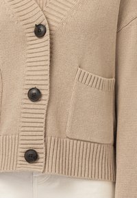 Beige stickad kofta med ribbade kanter, tre mörka knappar och en liten framficka över ljusfärgade byxor.