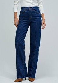 Mujer con jeans de cintura alta, de mezclilla azul oscura y corte recto, con costuras delanteras y tacones marrones de punta, contra un fondo liso.