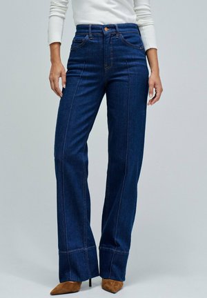 Mujer con jeans de cintura alta, de mezclilla azul oscura y corte recto, con costuras delanteras y tacones marrones de punta, contra un fondo liso.