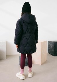 Mustast tikandiga sulejope kapuutsiga, vöökohas vöö, kombineeritud burgundia retuuside ja kreemjate tenniste ning roosade sokkidega. Tavaline taust.