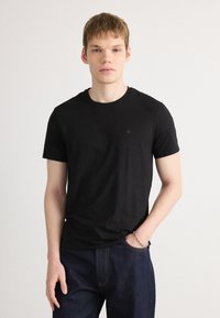 Black cotton t-shirt with short sleeves and a round neckline, featuring a small, subtle logo on the chest. Worn with dark denim jeans.