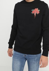 Sweatshirt nero realizzato in morbido misto cotone, con un design floreale rosso e verde sul lato sinistro del petto, polsini e vita a coste.