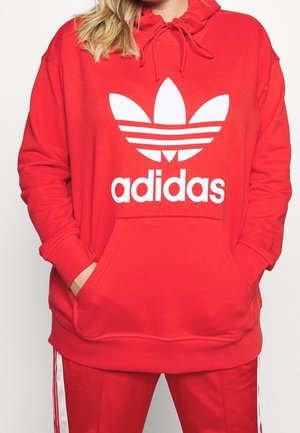 Osoba v červené mikině Adidas a červených kalhotách s bílými pruhy, ruce zasunuté v kapse mikiny.