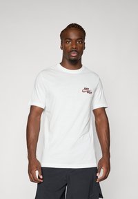 Hvid bomulds T-shirt med rund halsudskæring og korte ærmer, med en rød "JUST DO IT" logo på venstre brystområde.