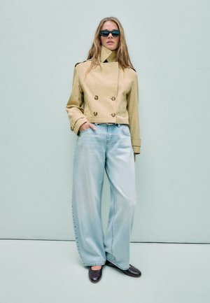 Ženska v bež skrajšanem plašču, širokih svetlo modrih kavbojkah, črnih čevljih in temnih sončnih očalih ob svetlo sivi steni.