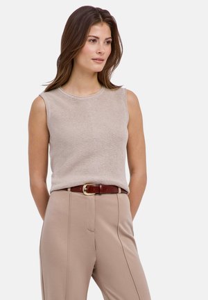Femme aux longs cheveux bruns portant un haut beige sans manches rentré dans un pantalon beige taille haute avec une ceinture marron, regardant sur le côté.