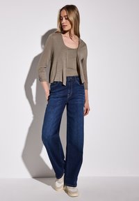 Ljusbeige öppen cardigan över en matchande tanktopp, parat med vida mörkblå jeans och krämfärgade sneakers. Slät tygkvalitet, avslappnad passform.