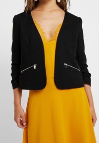 Black cropped jacket with ribbed texture, featuring two front zippered pockets. Worn over a yellow dress with lace neckline detailing.