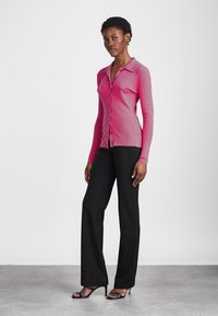 Cardigan rose à boutons avec col et manches longues, associé à un pantalon noir ajusté et des sandales à talons noires sur un sol gris.