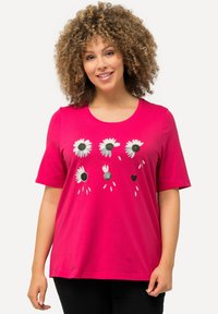 Camiseta de algodón rosa con mangas cortas, que presenta un gráfico floral de margaritas con centros negros y un pequeño diseño de corazón.