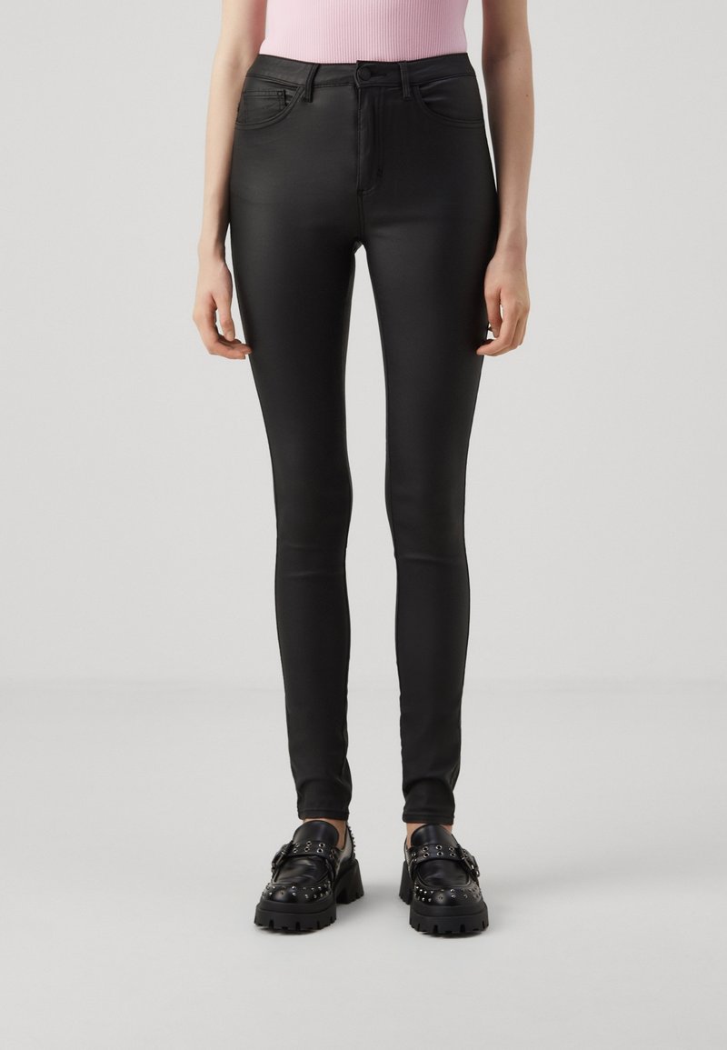 Femme portant un pantalon skinny taille haute noir avec des poches avant et des mocassins noirs cloutés épais, debout sur un fond uni.