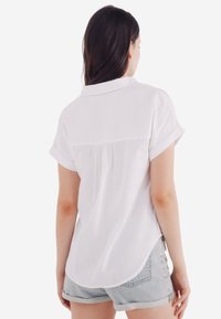 Witte korte mouwen knoopshirt met een ronde zoom, voorzien van een schouderpas en plooidetail op de rug, gedragen met denim shorts.