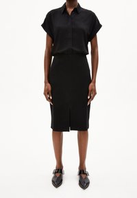 Blusa nera a maniche corte con colletto abbinata a una gonna a tubino nera al ginocchio con spacco frontale, completata da scarpe nere a punta.