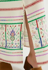 Jupe en tissu crème avec des motifs floraux et géométriques multicolores. Présente des fentes latérales et un ourlet large, complétée par des chaussures à talons en bois.