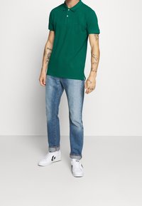 Polo vert avec col, manches courtes, et trois boutons, assorti à un jean bleu clair et des baskets blanches avec des accents noirs.