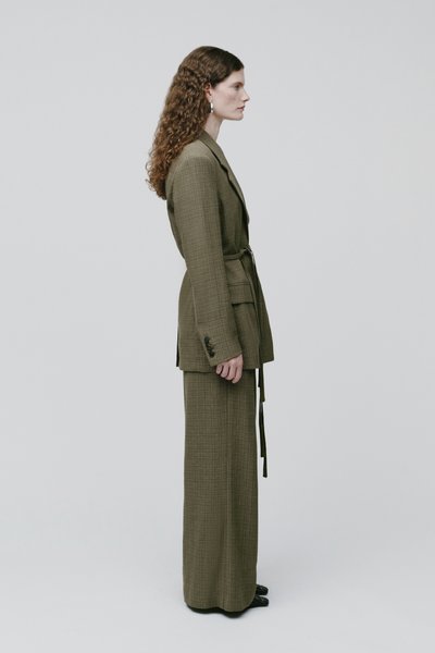 Femme aux cheveux bouclés portant un blazer ceinturé vert olive et un pantalon large assorti, debout de profil sur un fond uni.
