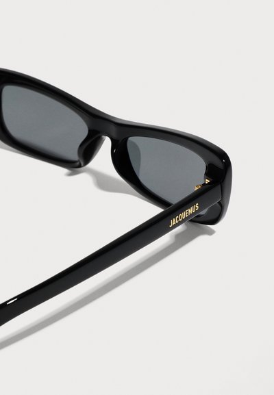 Lunettes de soleil rectangulaires noires avec verres foncés et "JACQUEMUS" écrit en or sur l'une des branches, sur un fond clair uni.