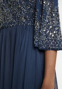 Vestido de noche azul marino con intrincados bordados de cuentas en el corsé, falda de tul fluida y mangas transparentes adornadas con lentejuelas y cuentas.
