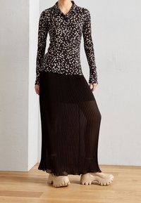 Blouse noire à manches longues avec des taches blanches, associée à une jupe plissée noire et des chaussures à plateforme beiges sur un sol en bois.