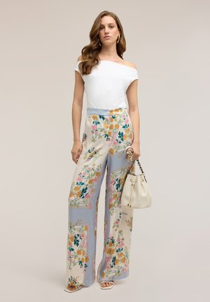 Kvinde med langt bølget hår iført hvid off-shoulder top, brede blomstrede bukser, hvide sandaler og holder en beige håndtaske med kædestrimmel.