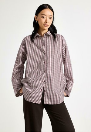 Donna con lunghi capelli neri che indossa orecchini a cerchio d'oro, una camicia a quadri rossa e bianca abbottonata e pantaloni neri, in piedi con le mani nelle tasche.