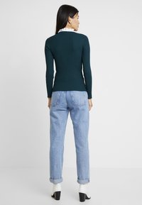 Donkergroene geribbelde trui met lange mouwen, voorzien van een witte kraag met ruches, gecombineerd met lichtblauwe hoog geplaatste spijkerbroek en witte laarzen met hak.