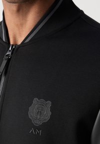 Gros plan d'une personne portant une veste zippée noire avec un visage de tigre subtilement embossé et un logo stylisé "AM" sur la poitrine.