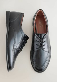 Miestne čierne lesklé kožené spoločenské topánky s brogue detailmi a čiernymi šnúrkami, zobrazené z vrchu na svetlej ploche.