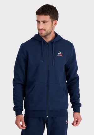 Námořnická modrá mikina s kapucí na zip vyrobená z měkké tkaniny, s nastavitelnou kapucí, předními kapsami a malým logem na hrudi.