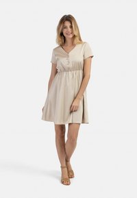 Korte mouwen, beige jurk met een V-hals, een geraffineerde taille en knoopdetails. Het soepele materiaal benadrukt de vloeiende rok. Gecombineerd met bruine sandalen met hakken.
