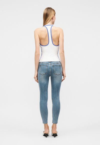 Γυναίκα που φοράει λευκή αμάνικη μπλούζα με ρακερ πλάτη και μπλε λεπτομέρειες, ανοιχτό μπλε στενά τζιν και μαύρα παπούτσια με ψηλό τακούνι, γυρισμένη προς τα πίσω σε απλό φόντο.