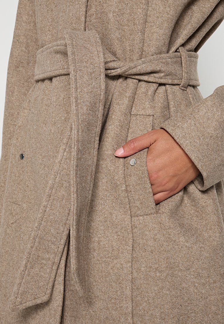 Abrigo de lana beige con cintura ceñida, bolsillos laterales y una textura suave, con detalles de botones grises cerca de los bolsillos.
