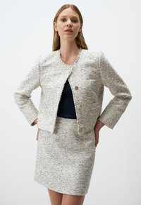 Giacca in tweed bianco e blu navy con bordi sfrangiati, scollo rotondo e due bottoni dorati. Abbinata a una gonna in tweed coordinata.