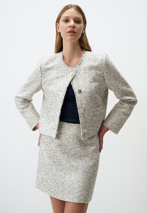 Veste en tweed blanc et marine avec des bords effilochés, un col rond et deux boutons dorés. Associée à une jupe en tweed assortie.