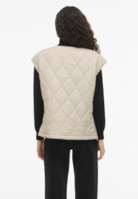 Tikanditega vattitud varrukateta vest helebeezhis toonis, millel on ruutmustriga tikand, kantud musta pika varrukaga topi peal. Otse lõikejoonega allserv ja mugav lõige.