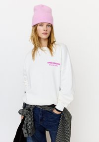 White sweatshirt with pink "APRÈS SEASON" text, paired with blue jeans. A black and white checkered shirt is tied around the waist. Pink knit beanie.