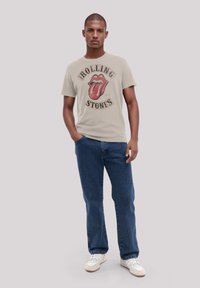 Beige cotton T-shirt featuring a graphic of red lips and text "ROLLING STONES." Paired with blue denim jeans and white sneakers.