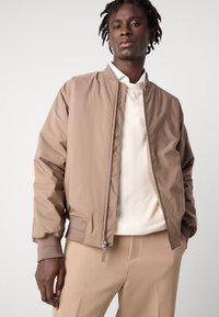 Beige bomberjack met een gladde textuur, ribgebreide manchetten en een ritssluiting aan de voorkant. Draag het over een lichtcrème shirt, gecombineerd met beige broek.