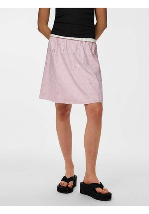 Kobieta nosząca jasnoróżową kwiatową spódnicę z oczkami, czarny top i czarne klapki na platformie, stoi z rękami za plecami.