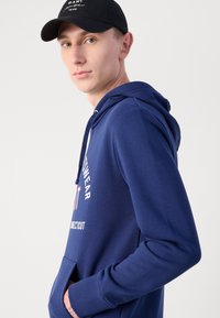 Sudadera con capucha azul marino con bolsillo frontal, con texto y logotipo en blanco en el pecho. Tejido de mezcla de algodón, textura suave.