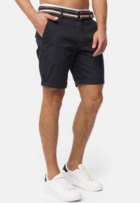 Shorts en coton bleu marine avec des revers et des poches latérales, associés à une ceinture multicolore à rayures et des tennis blanches avec des accents noirs.