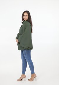 Olivgrön, flödande ytterplagg med hög krage och vida ärmar, matchat med blå jeans och klara klacksandaler. Slät textur, minimalistisk design.