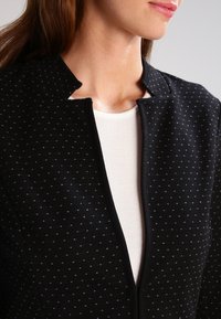 Černá bunda s texturovanou tkaninou a malými bílými trojúhelníkovými vzory. Má minimalistický límec a přední zip. Nositá přes bílou košili.