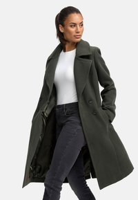 Dark green wool coat with a double-breasted design, notched collar, and side pockets. Lined interior, worn over a white top and black jeans.